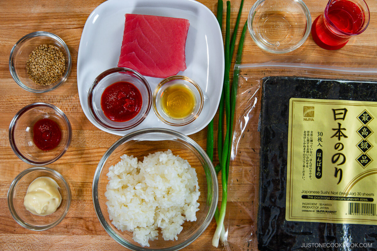 A top-down view of sushi ingredients on a wooden surface: a block of raw tuna, sushi rice, nori sheets, green onions, sesame seeds, sauces in small bowls, and mayonnaise—perfect for making a spicy tuna roll.