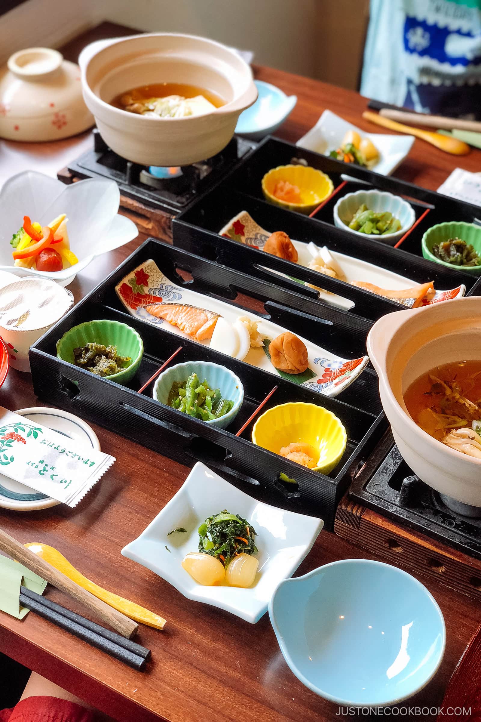 A table set with a traditional Japanese meal, featuring colorful dishes in bento trays and bowls, beautifully arranged according to Japanese dining etiquette. Vegetables, pickles, fish, and two hot pots complete the authentic experience.