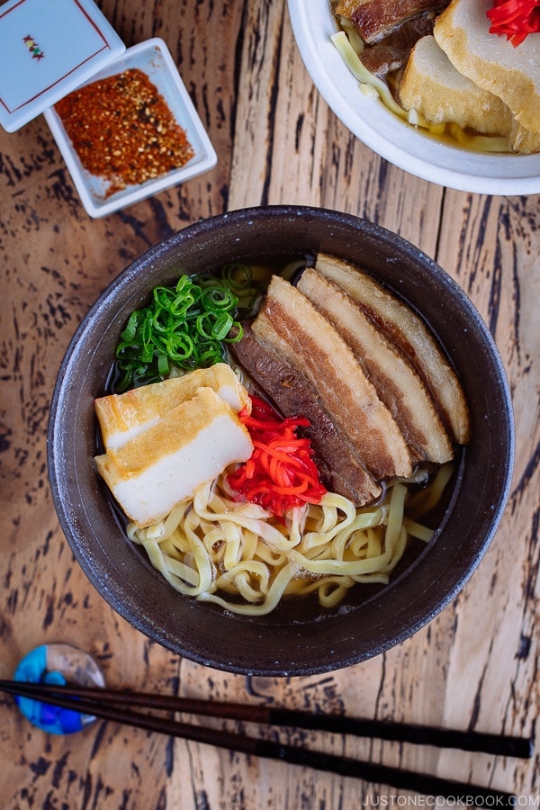 A white bowl containing Okinawa Soba and delicious Japanese dashi and pork broth, topped braised pork belly, fish cakes, green onion, and red pickled ginger.