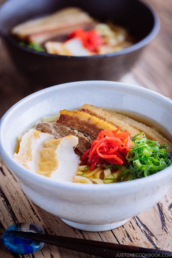 A white bowl containing Okinawa Soba and delicious Japanese dashi and pork broth, topped braised pork belly, fish cakes, green onion, and red pickled ginger.