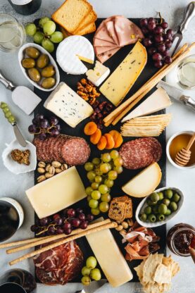 Cheese board filled with cheese, cold cuts, dried fruits, bread sticks, olives, grapes, and condiments.