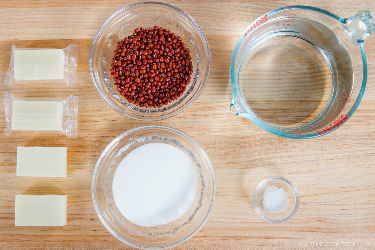 A top-down view of ingredients for zenzai on a wooden surface: three blocks of butter, two bowls with red beans and sugar, a glass measuring cup with water, and a small bowl of salt.