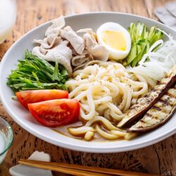 A white bowl containing cold curry udon with sliced pork, boiled egg, and fresh vegetables.