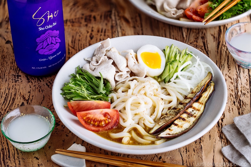 A white bowl containing cold curry udon with sliced pork, boiled egg, and fresh vegetables.