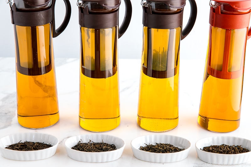 different types of hojicha tea leaves in white dish in front of pitchers of tea