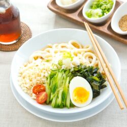 A white bowl containing chilled udon noodles, tenkasu, julienned cucumber, boiled egg, wakame seaweed, and grated onion, along with savory noodle soup.