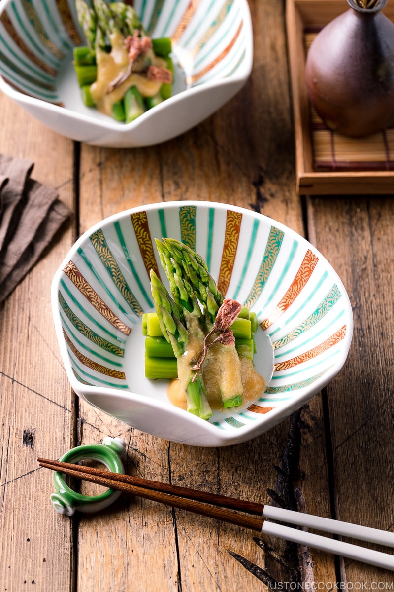 A shell shaped bowl containing Asparagus with Miso Dressing (Karashi Sumisoae) garnished with salt pickled cherry blossom.