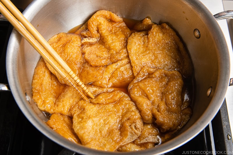 A stainless steel pot filled with simmering, golden-brown inari sushi tofu pouches soaked in broth. Wooden chopsticks rest on the side, ready to pick up a savory tofu piece.