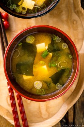 A bowl of miso soup, made from an authentic miso soup recipe with tofu cubes, seaweed, and sliced green onions, served on a wooden tray with red chopsticks beside it.