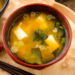 A bowl of miso soup with tofu cubes, seaweed, and chopped green onions sits on a wooden tray next to chopsticks and a red flower-shaped dish—a classic presentation for any authentic miso soup recipe.