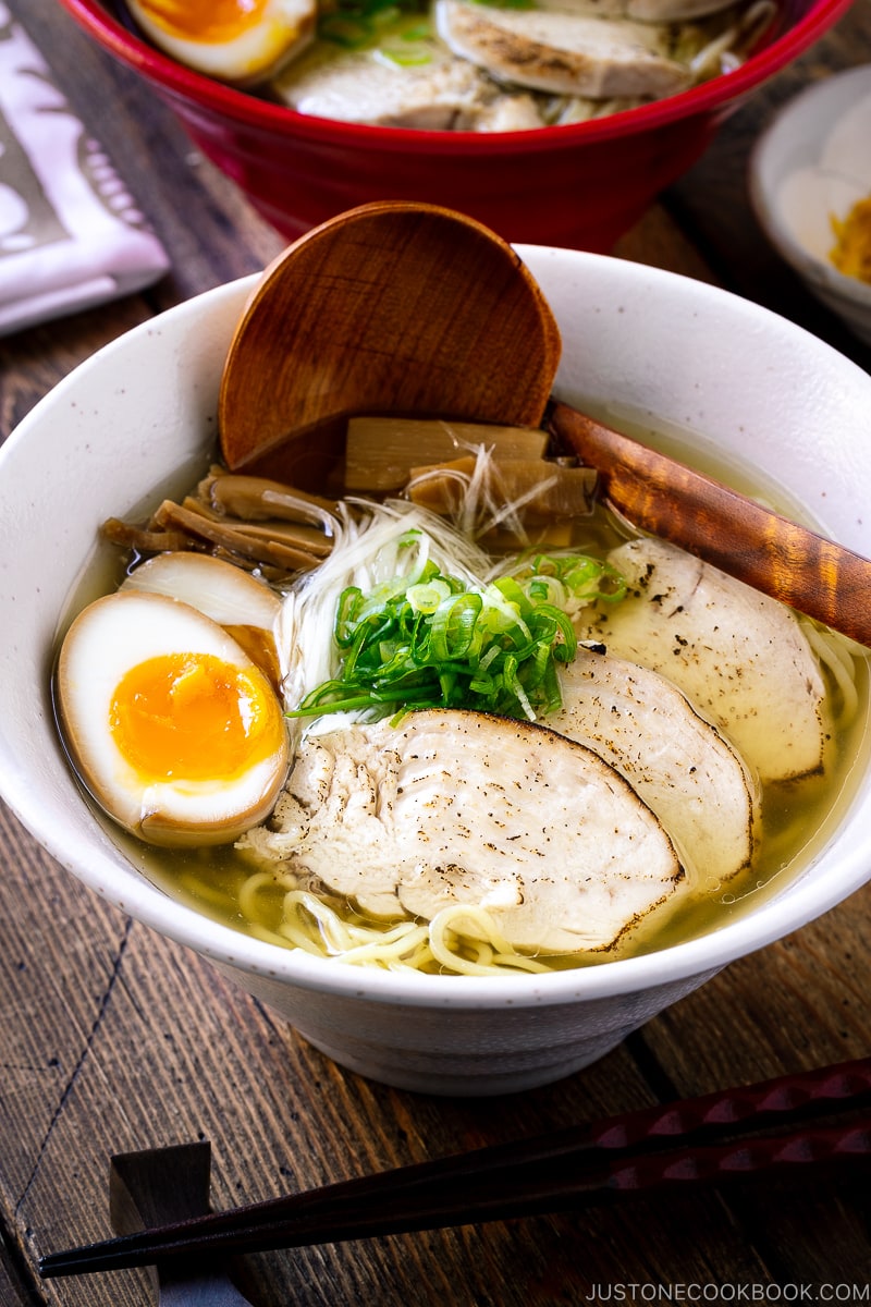 A white bowl containing Shio Ramen, topped with Chicken Chashu, Ramen Egg, bamboo shoot, white negi, and green onion.