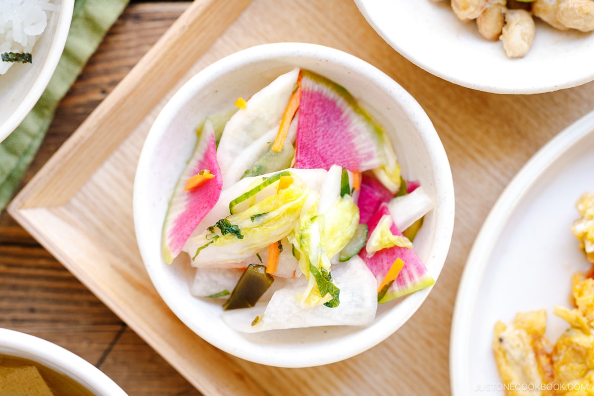 A small white bowl filled with vibrant Japanese pickles, including slices of radish, napa cabbage, and carrots, served on a wooden tray.
