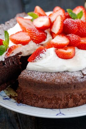 A large plate containing Chocolate Gateau (Chocolate Cake) dusted with powdered sugar and decorated with whipped cream and strawberries.