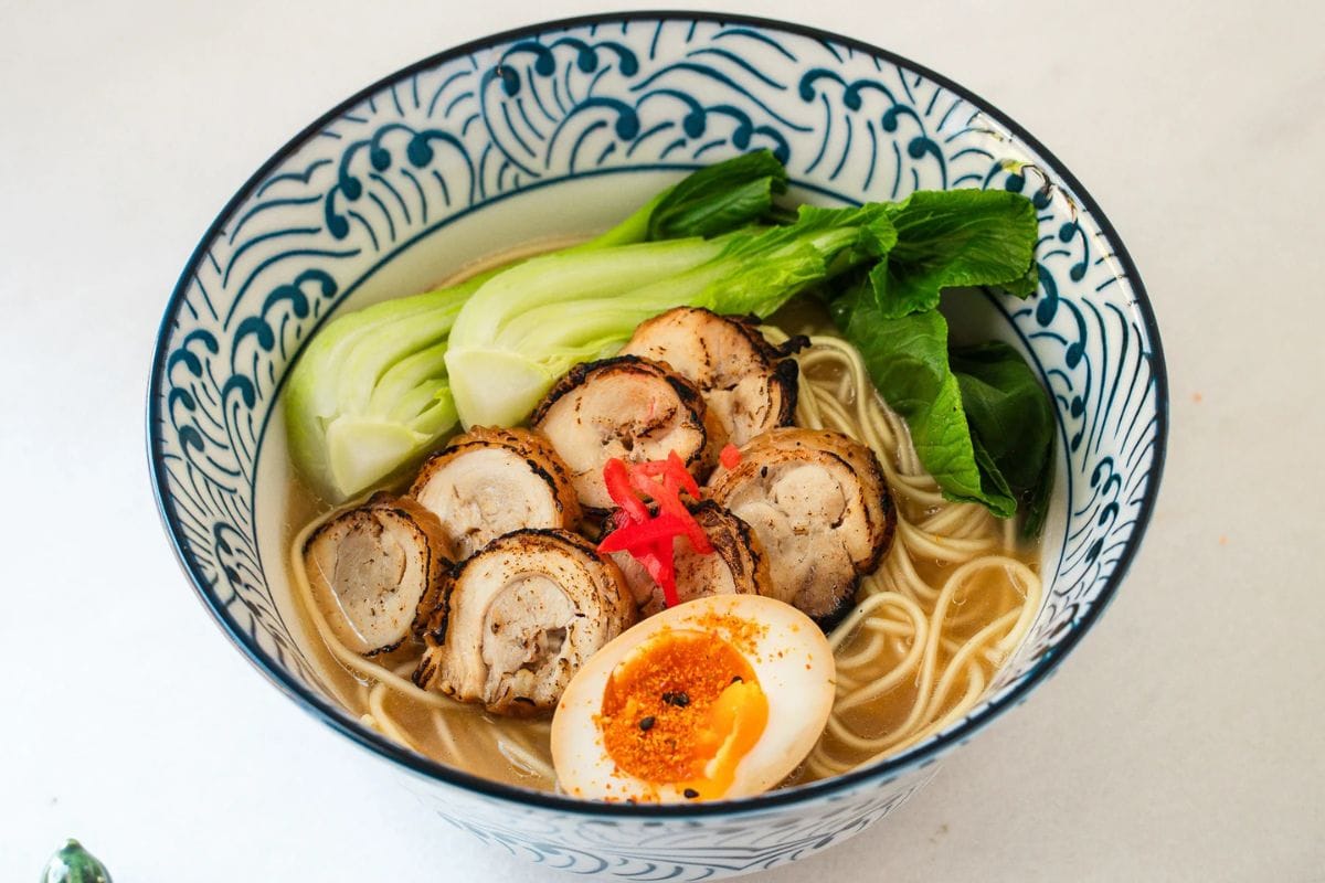 Juicy ramen bowl with sliced pork, soft-boiled egg, fresh vegetables, and flavorful broth in a decorative blue and white bowl.