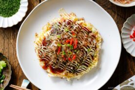 A white ceramic plate containing Okonomiyaki, a savory Japanese cabbage pancake topped with okonomi sauce, Kewpie mayo, and bonito flakes.