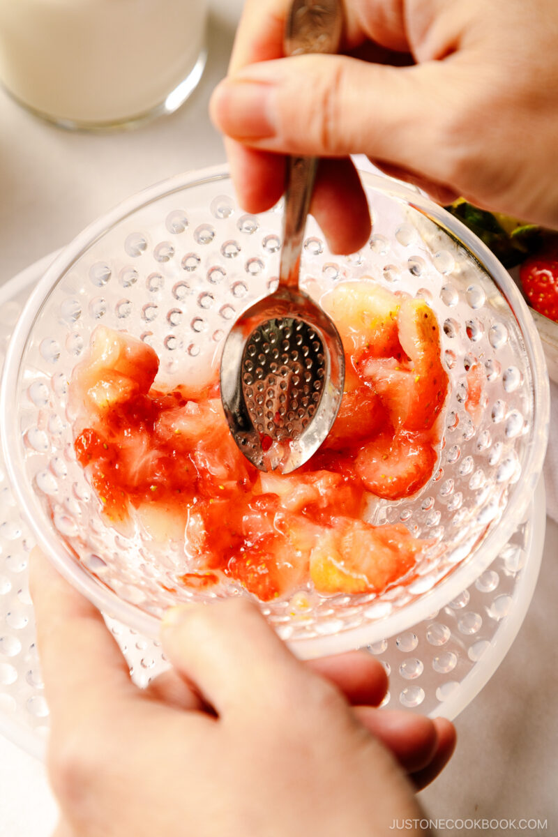 Strawberries in a glass bowl are being mashed by the spoon.
