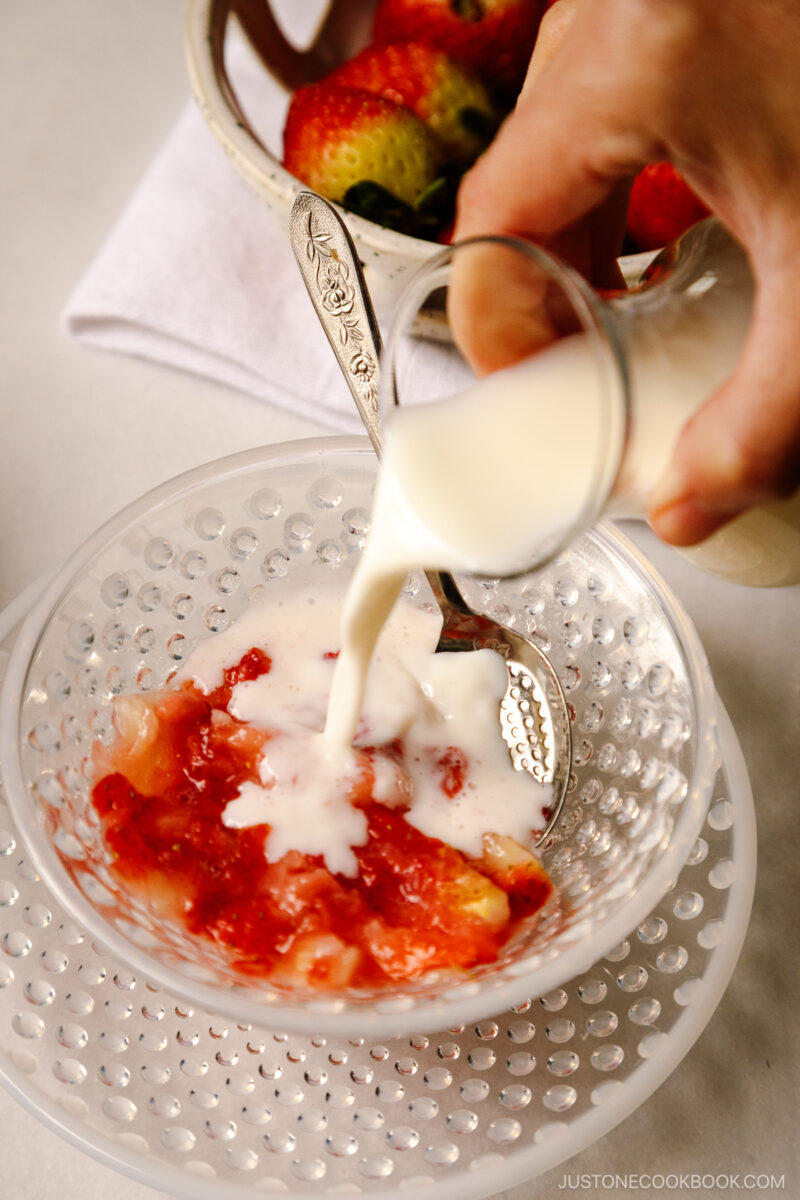 Milk in a glass bottle is being pored into the mashed strawberries in a glass bowl.