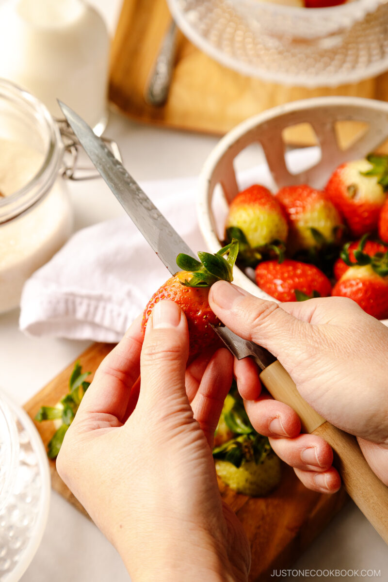 The green leaf on top of the strawberries is being removed by a sharp knife.