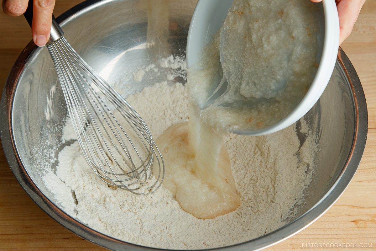 A person pours a bowl of wet mixture into a large metal bowl of flour mixture while holding a whisk, preparing to mix ingredients for homemade okonomiyaki on a wooden surface.