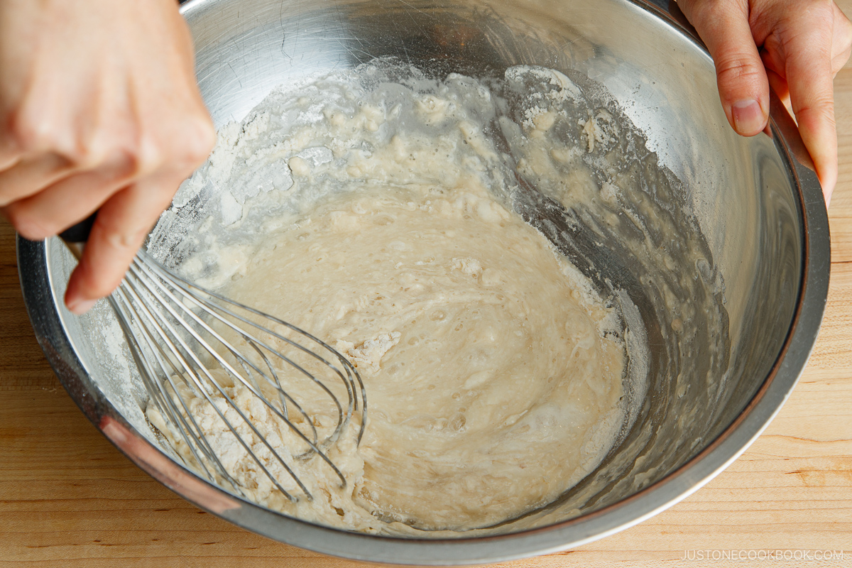 A person uses a metal whisk to mix okonomiyaki dough in a large metal bowl on a wooden surface.