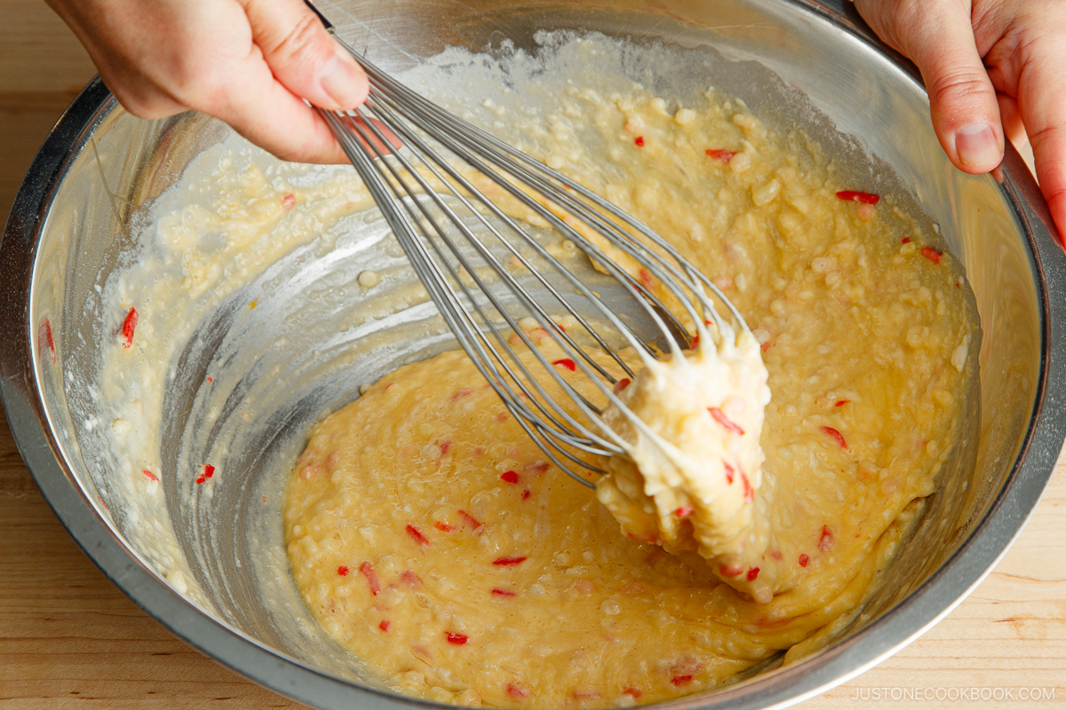 Close-up of hands using a whisk to mix thick okonomiyaki batter with visible red pieces in a large metal bowl on a wooden surface.