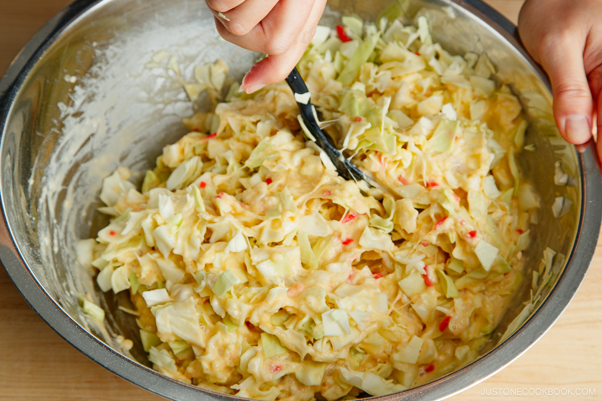 A person mixing chopped cabbage, mayonnaise, and other ingredients in a large metal bowl with a black spatula, preparing a creamy salad or an okonomiyaki-inspired coleslaw.
