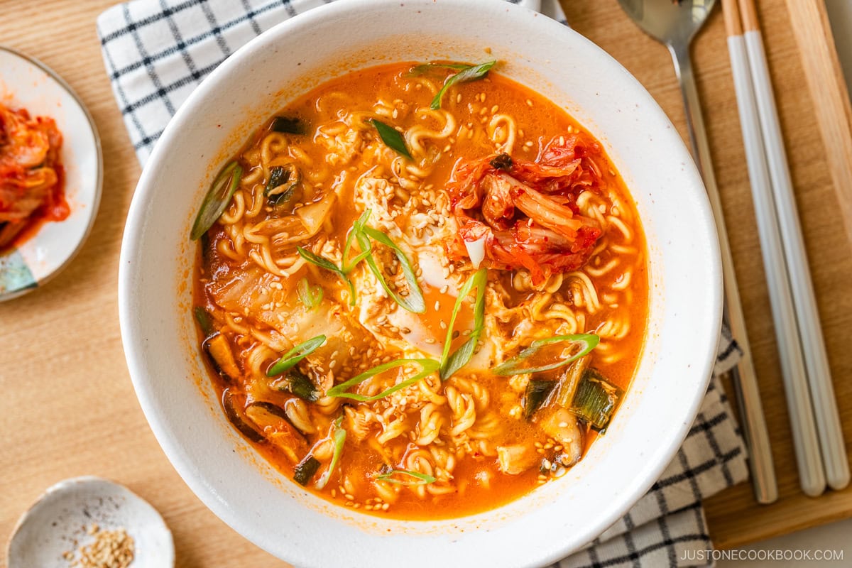 A white ramen bowl containing Korean ramen (ramyeon), topped with soft-cooked egg, kimchi, and green onions.