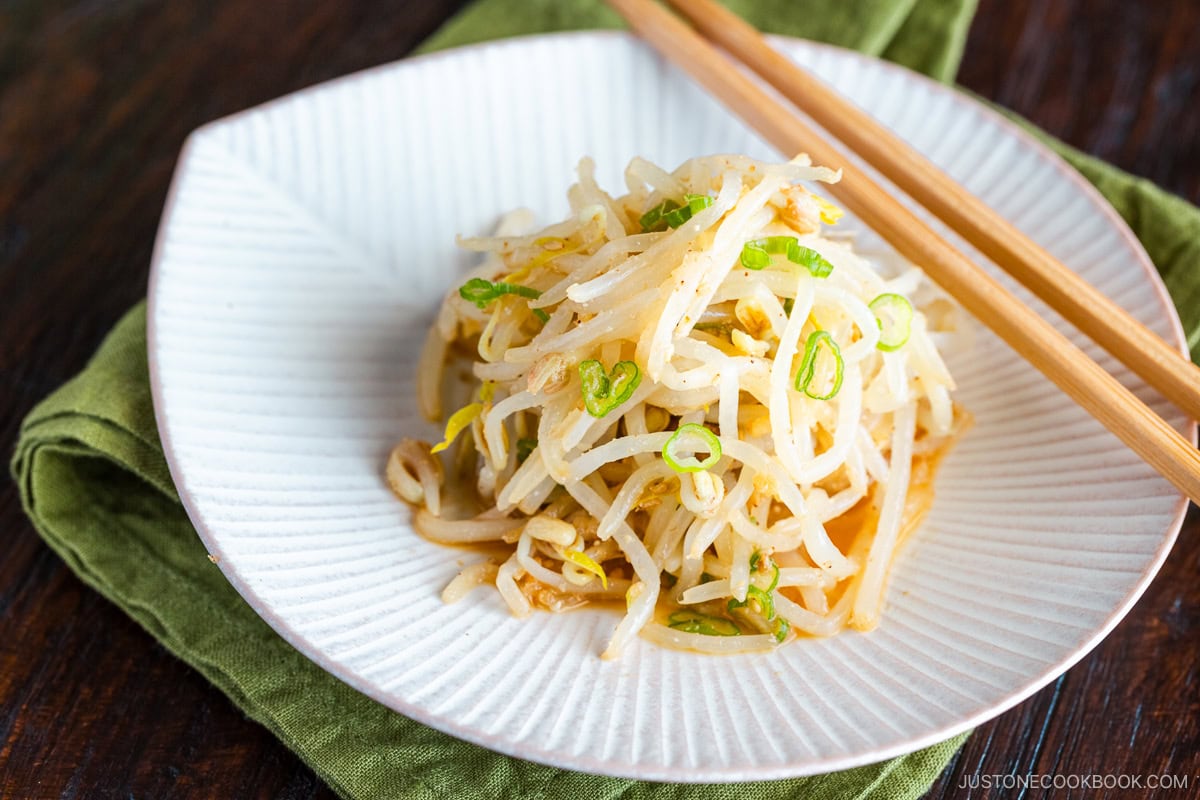 A white plate containing Bean Sprout Salad, seasoned with sesame oil and mixed with (ground) sesame seeds and green onions.