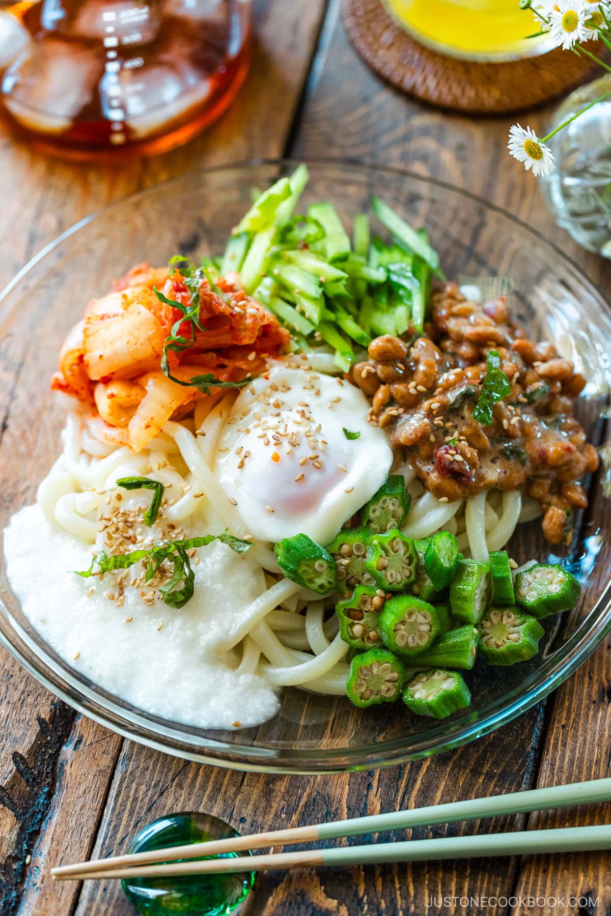 A glass plate containing cold natto udon topped with kimchi, onsen tamago, blanched okra, fresh cucumber, and grated nagaimo.