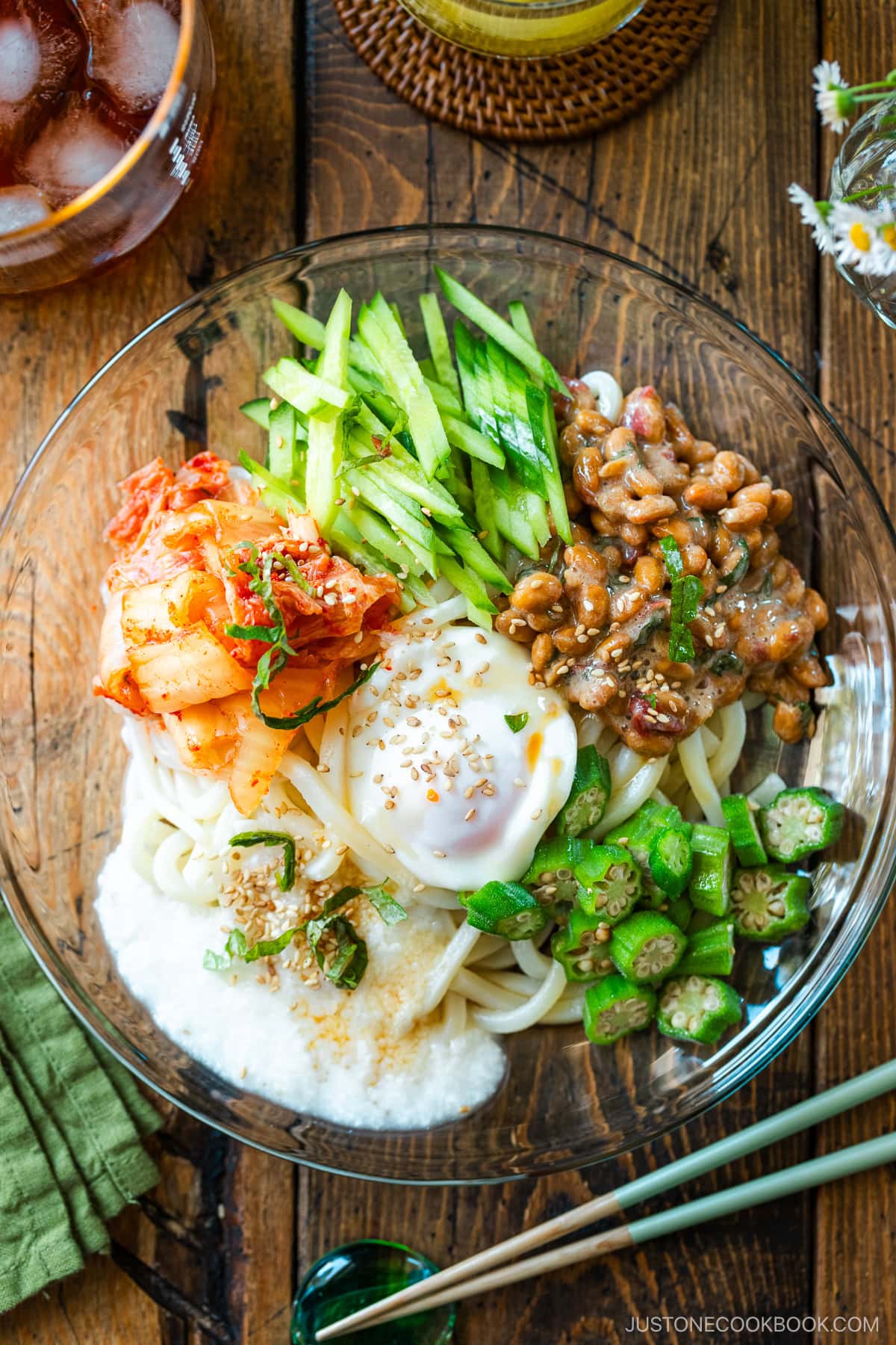 A glass plate containing cold natto udon topped with kimchi, onsen tamago, blanched okra, fresh cucumber, and grated nagaimo.