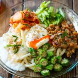 A glass plate containing cold natto udon topped with kimchi, onsen tamago, blanched okra, fresh cucumber, and grated nagaimo.
