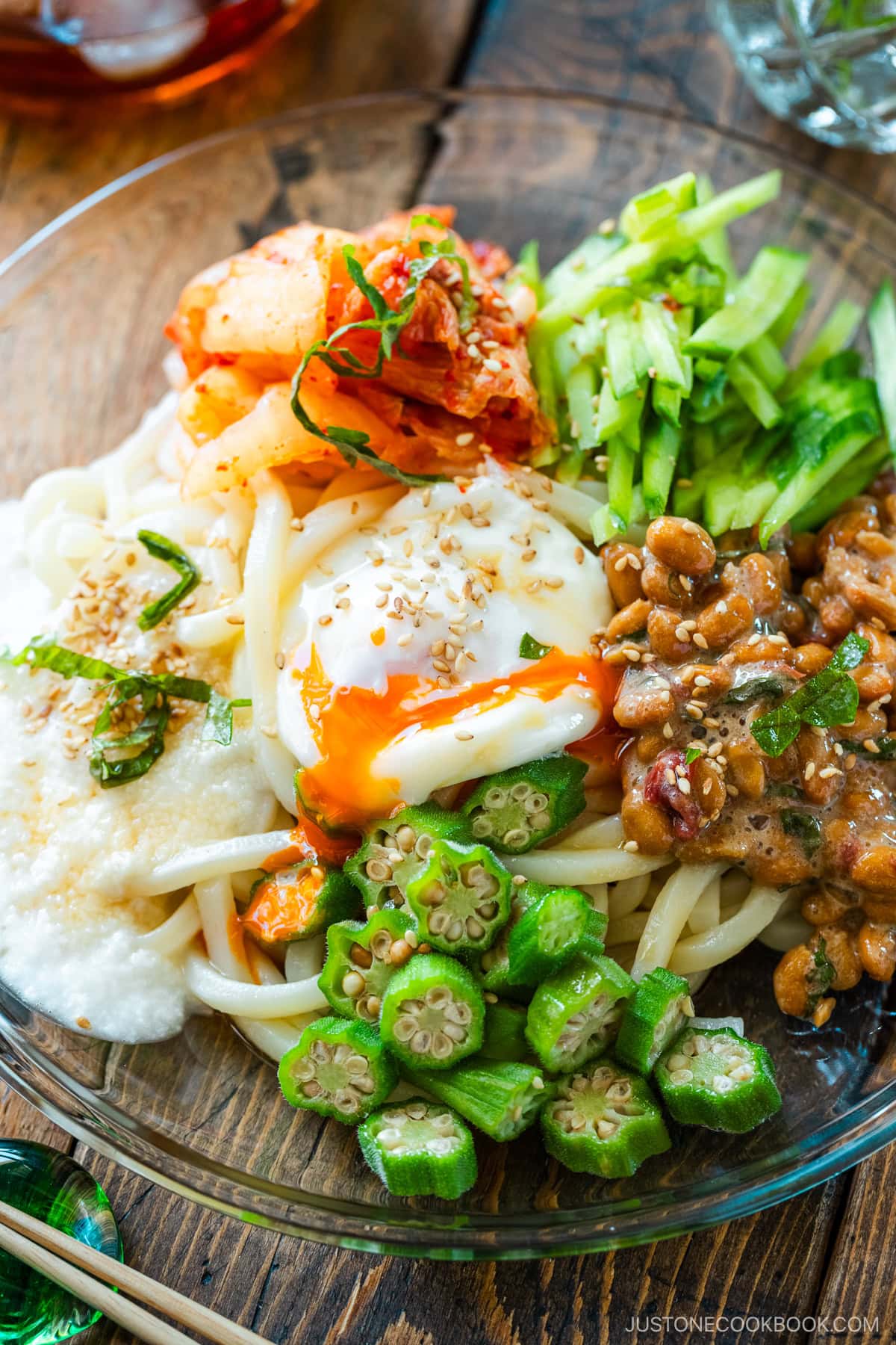 A glass plate containing cold natto udon topped with kimchi, onsen tamago, blanched okra, fresh cucumber, and grated nagaimo.