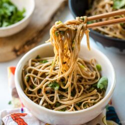 Chopsticks pulling noodles from a white bowl containing Soba Noodle Salad topped with sesame seeds and chopped cilantro and green onions.