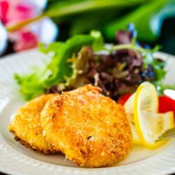 A white plate containing creamy crab croquettes, served with salad and a slice of lemon.