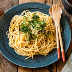 A black round plate containing metaiko pasta - Japanese spaghetti with spicy salted cod roe topped with julienned shiso and shredded nori seaweed.