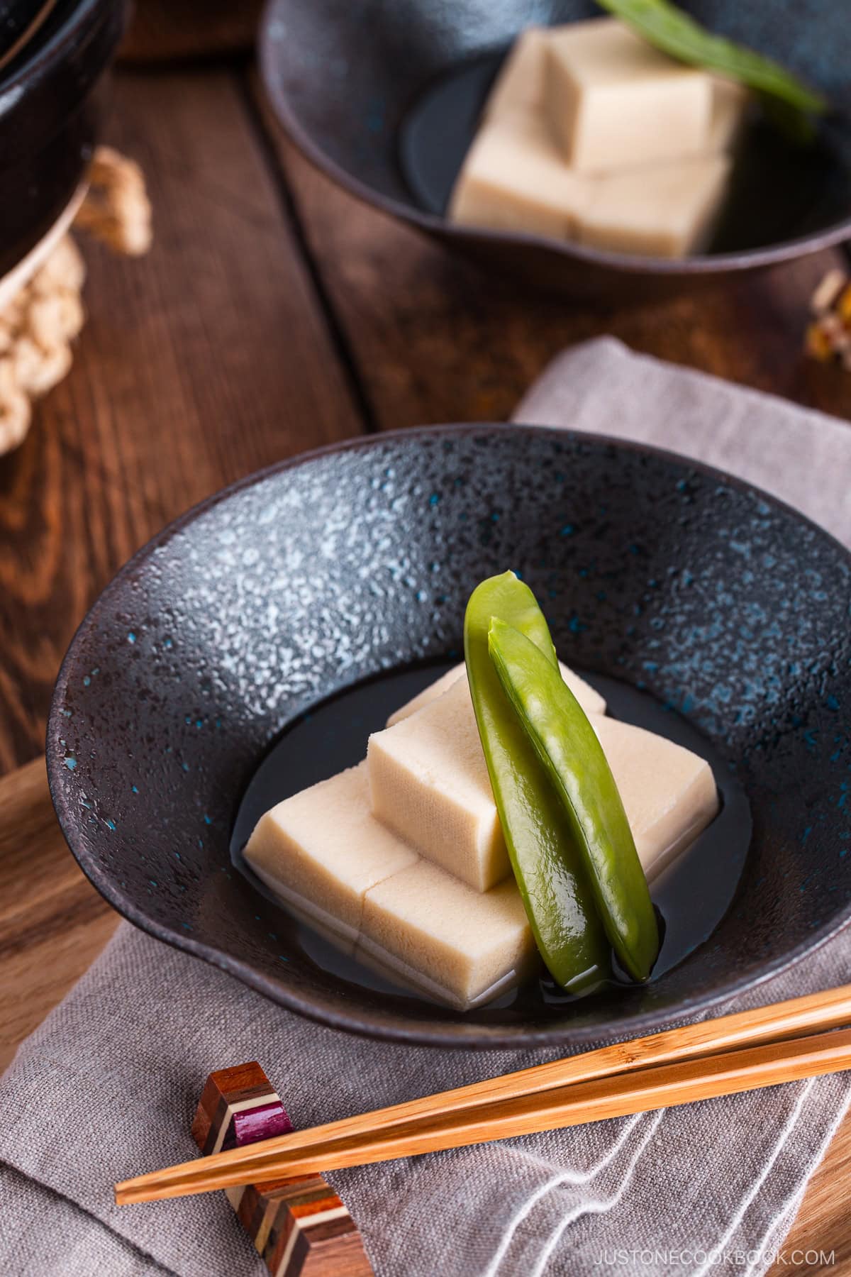Simmered koyadofu with snow peas in a black ceramic bowl.