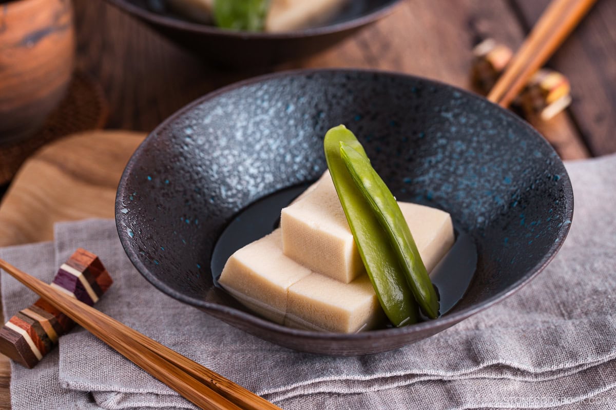 Simmered koyadofu with snow peas in a black ceramic bowl.