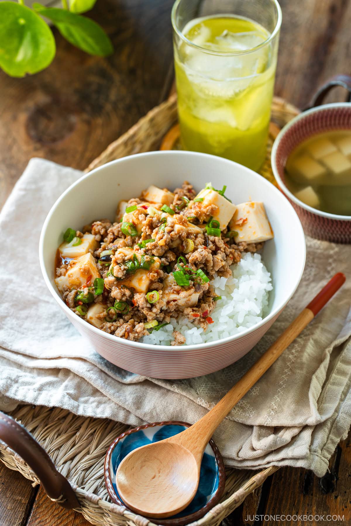 A donburi bowl containing mapo tofu over bed of steamed rice, served with a small bowl of miso soup and a glass of green tea.