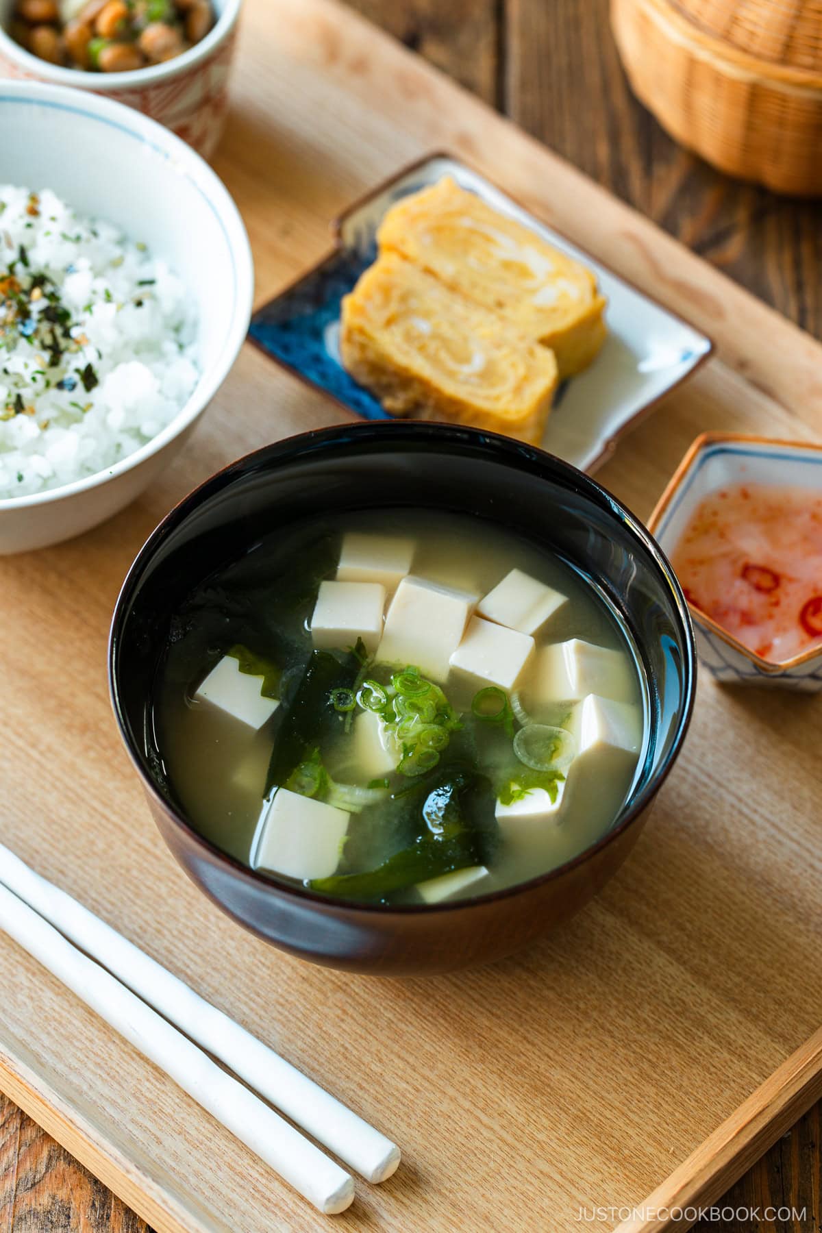 A light-colored wooden tray containing a bowl of miso soup with soft tofu, wakame seaweed, and thinly sliced green onions, along with a bowl of steamed rice, a small plate of tamagoyaki, and a smalll side dish.