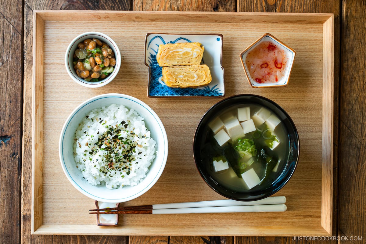 A light-colored wooden tray containing a bowl of miso soup with soft tofu, wakame seaweed, and thinly sliced green onions, along with a bowl of steamed rice, a small plate of tamagoyaki, and a smalll side dish.