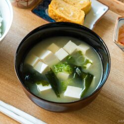 A light-colored wooden tray containing a bowl of miso soup with soft tofu, wakame seaweed, and thinly sliced green onions, along with a bowl of steamed rice, a small plate of tamagoyaki, and a smalll side dish.