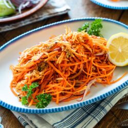An oval scalloped plate containing shredded carrot and tuna salad garnished with parsley and lemon.