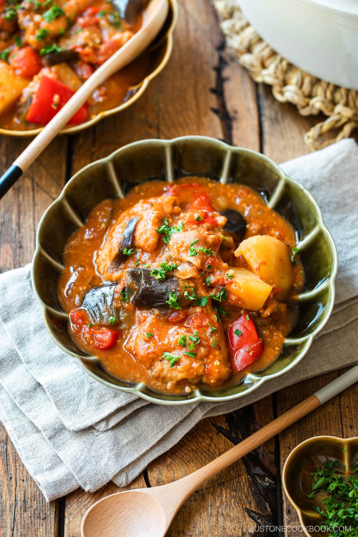 Fluted bowls containing Miso Tomato Chicken Stew garnished with chopped parsley on top, served with a glass of tea, a small dish of parsley, and a pot of the stew on the wooden table.