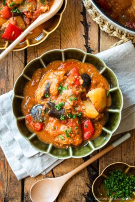Fluted bowls containing Miso Tomato Chicken Stew garnished with chopped parsley on top, served with a glass of tea, a small dish of parsley, and a pot of the stew on the wooden table.