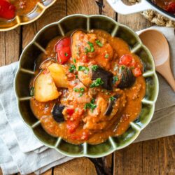Fluted bowls containing Miso Tomato Chicken Stew garnished with chopped parsley on top, served with a glass of tea, a small dish of parsley, and a pot of the stew on the wooden table.