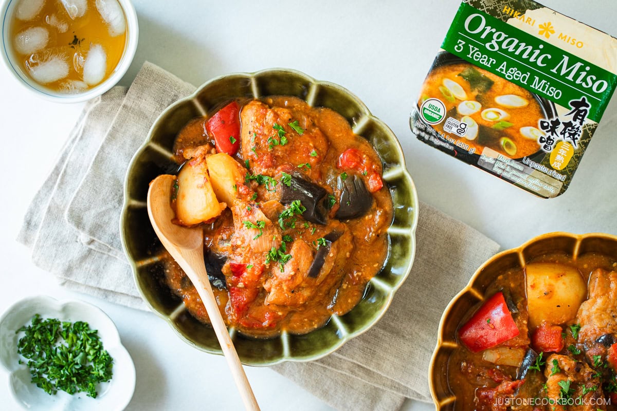 Fluted bowls containing Miso Tomato Chicken Stew garnished with chopped parsley on top, served with a glass of tea, a small dish of parsley, and a pot of the stew on the wooden table.