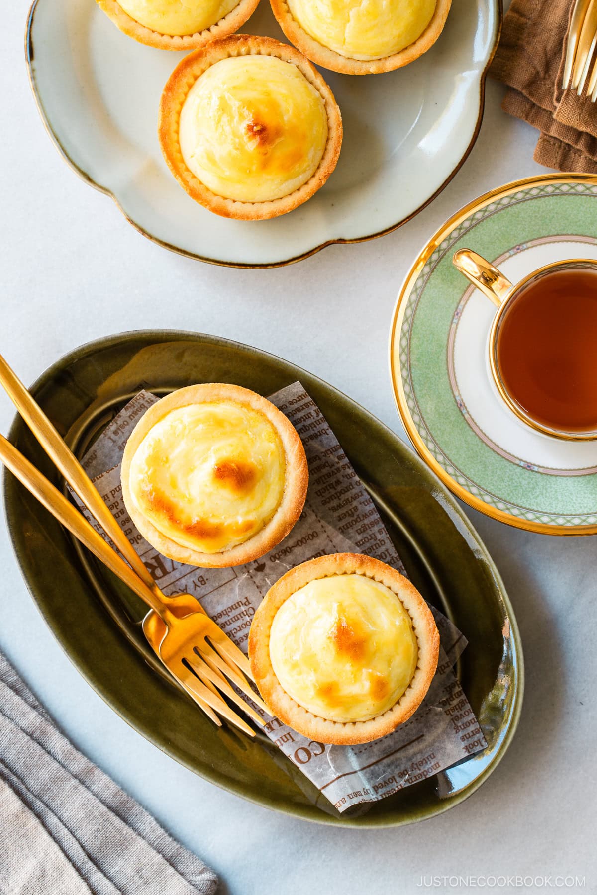 An olive oval plate and a white flower-shaped plate containing baked cheese tarts, served with a cup of tea.