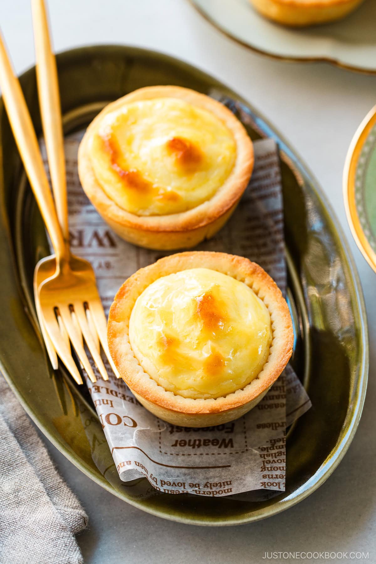 An olive oval plate and a white flower-shaped plate containing baked cheese tarts, served with a cup of tea.