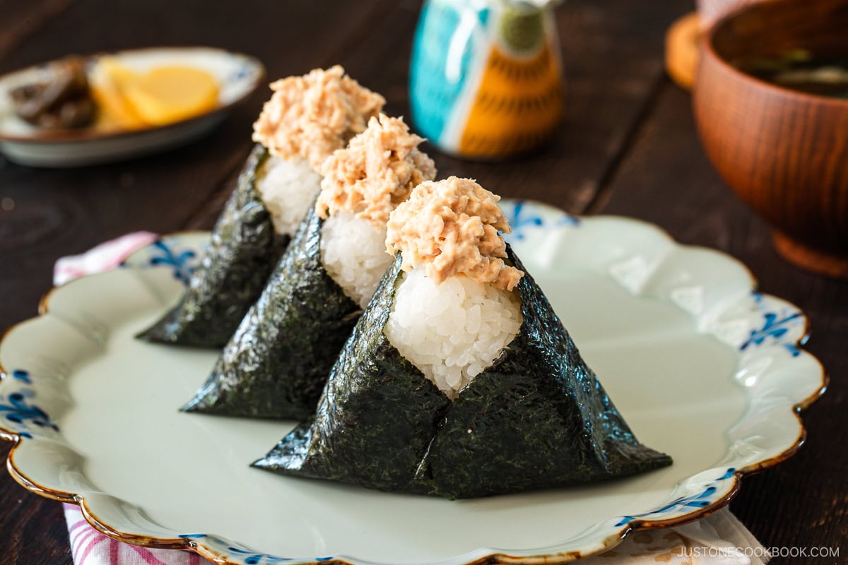 A plate containing three tuna mayo onigiri served with a wooden bowl of miso soup, a cup of tea, and a small dish of pickles.