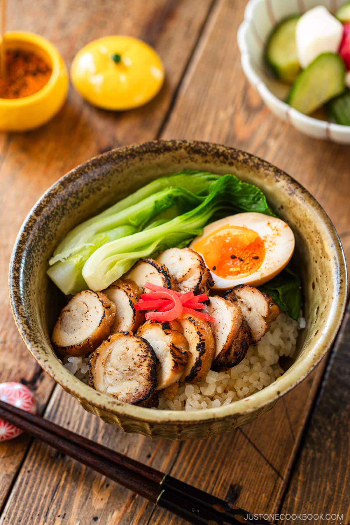A donburi bowl containing steamed rice topped with seared sliced chicken chashu, soft boiled egg, and blanched bok choy, garnished with pickled ginger.